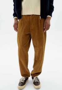 Braune Cordhosen mit vertikaler Rippung, die eine lockere Passform und elastische Bündchen aufweisen, kombiniert mit dunklen Schuhen und einem hellen Pullover.