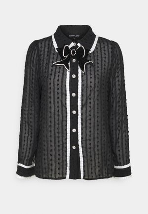 Blouse noire transparente avec des rayures verticales texturées, bordure blanche à volants le long de la patte de boutonnage et des poignets, et un nœud orné de bijoux au col.