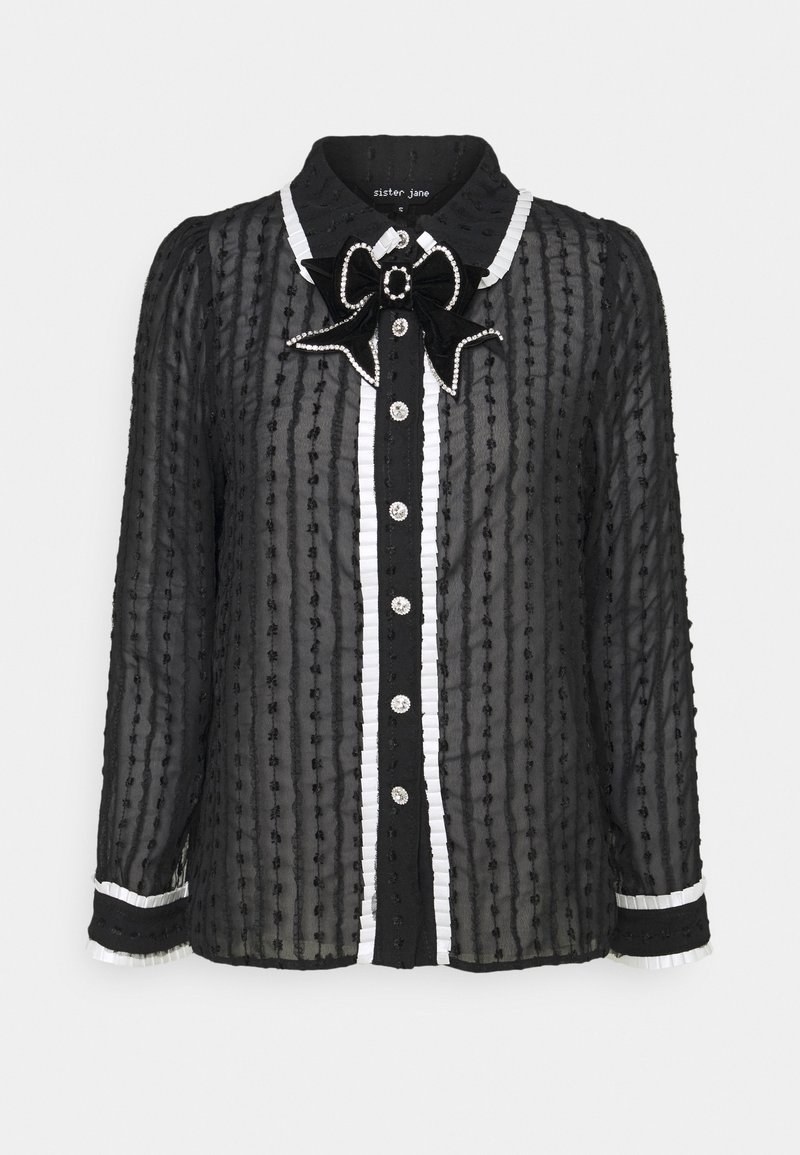 Blouse noire transparente avec des rayures verticales texturées, bordure blanche à volants le long de la patte de boutonnage et des poignets, et un nœud orné de bijoux au col.