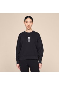 Black sweatshirt with a relaxed fit, round neckline, and dropped shoulders featuring a white logo on the chest. Smooth fabric texture.