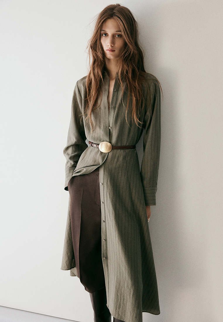 Femme aux longs cheveux bruns porte une robe boutonnée verte olive ceinturée sur un pantalon marron foncé, debout contre un mur clair uni.