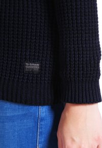 Černý pletený svetr s texturovaným vzorem, na přední levé straně s nášivkou G-Star. Hladké manžety a lem, nošený přes modré džíny.