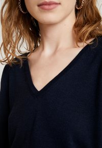 Granatowy sweter w serek wykonany z miękkiego materiału; charakteryzuje się teksturowanym wykończeniem. Godnym uwagi detalem jest delikatne przeszycie dekoltu.