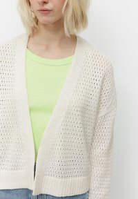 Cardigan bianco a maglia aperta con design a V indossato sopra una camicia aderente di colore verde chiaro. Il cardigan presenta un motivo testurizzato e polsini a costine.