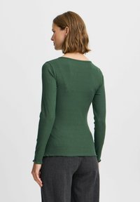 Grünes, geripptes Langarmoberteil mit rundem Ausschnitt, das sich durch ein tailliertes Design und eine leichte Textur auszeichnet, kombiniert mit dunkelgrauen Hosen.