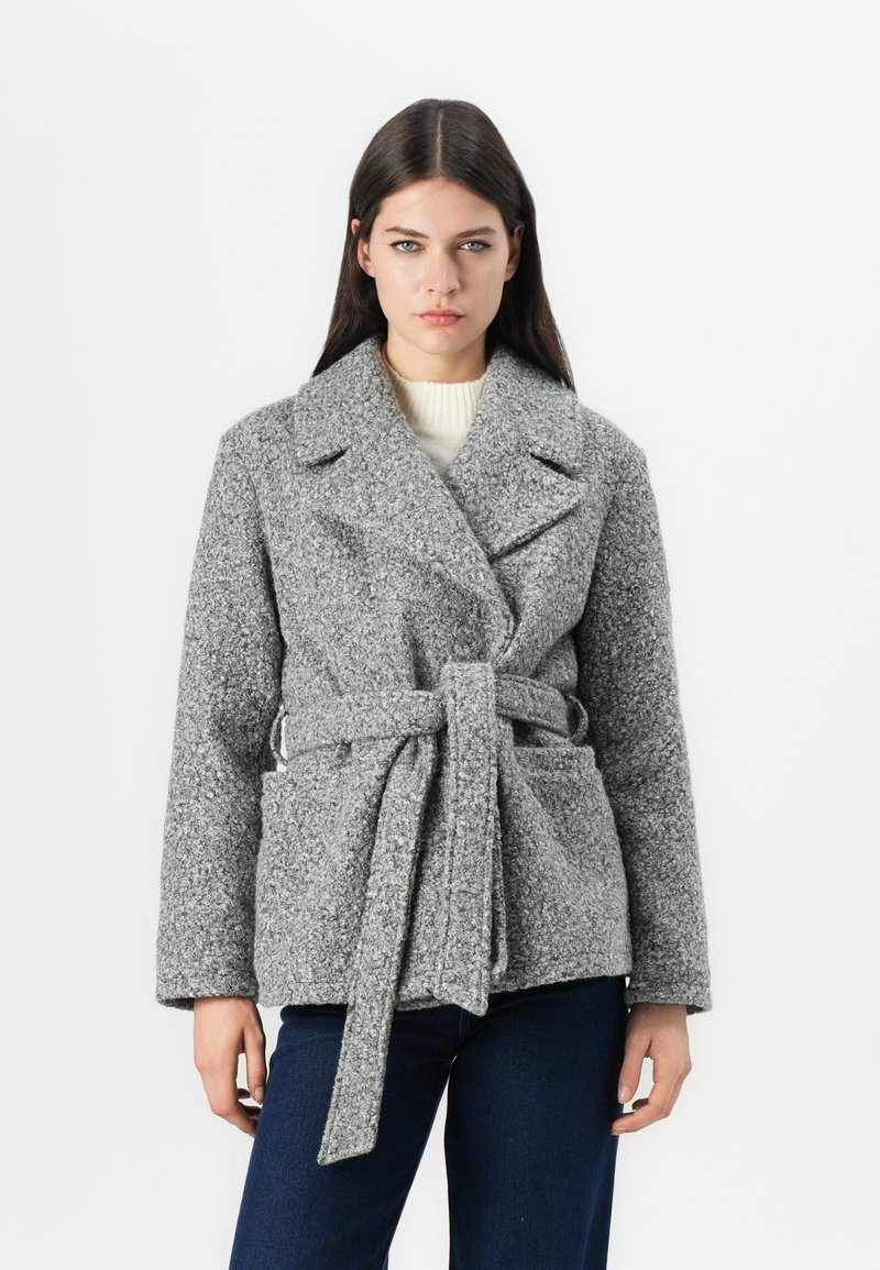 Grå tekstureret frakke med bælte, brede klapper og to forlommer, parret med en lys sweater under.