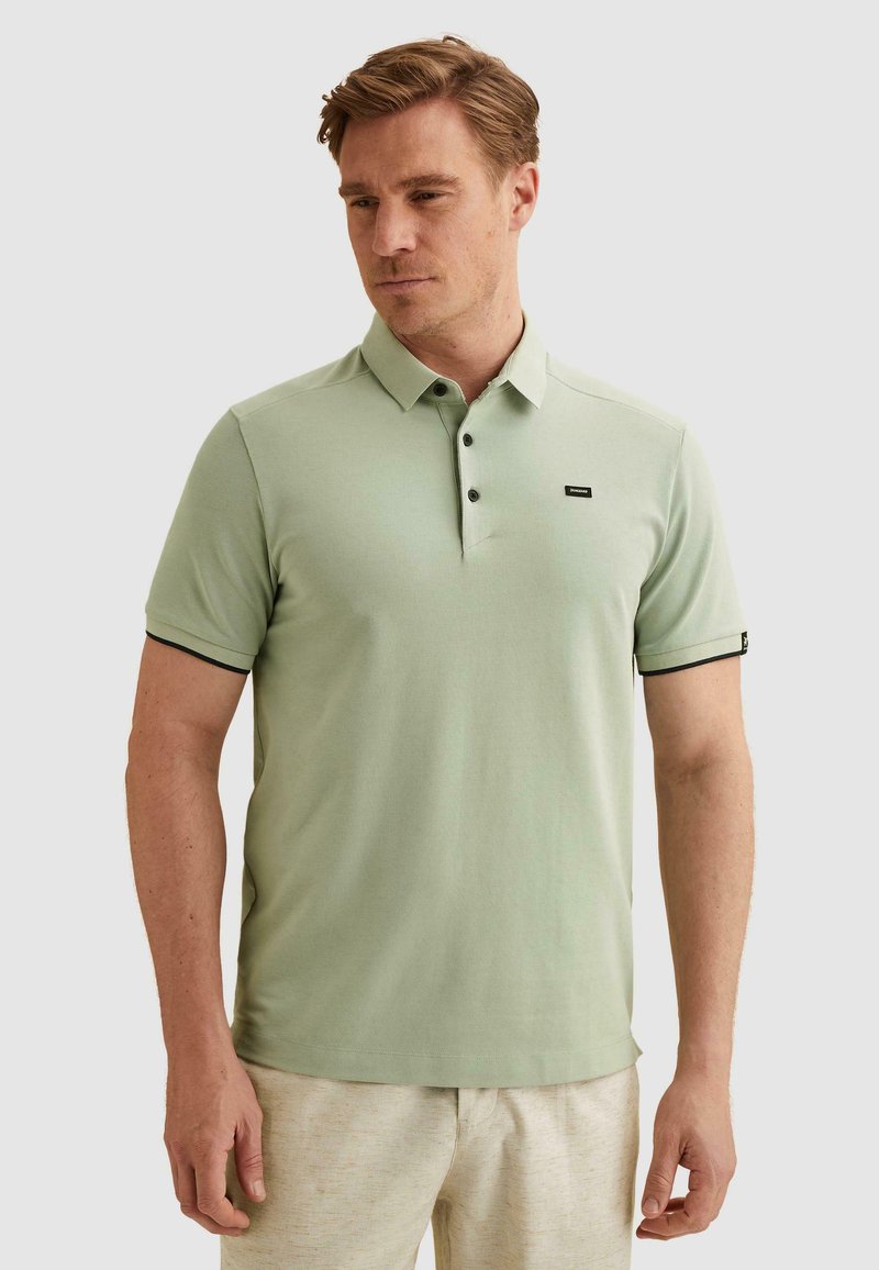 Homme portant un polo vert clair à manches courtes avec des boutons et un pantalon beige, debout devant un fond clair uni.