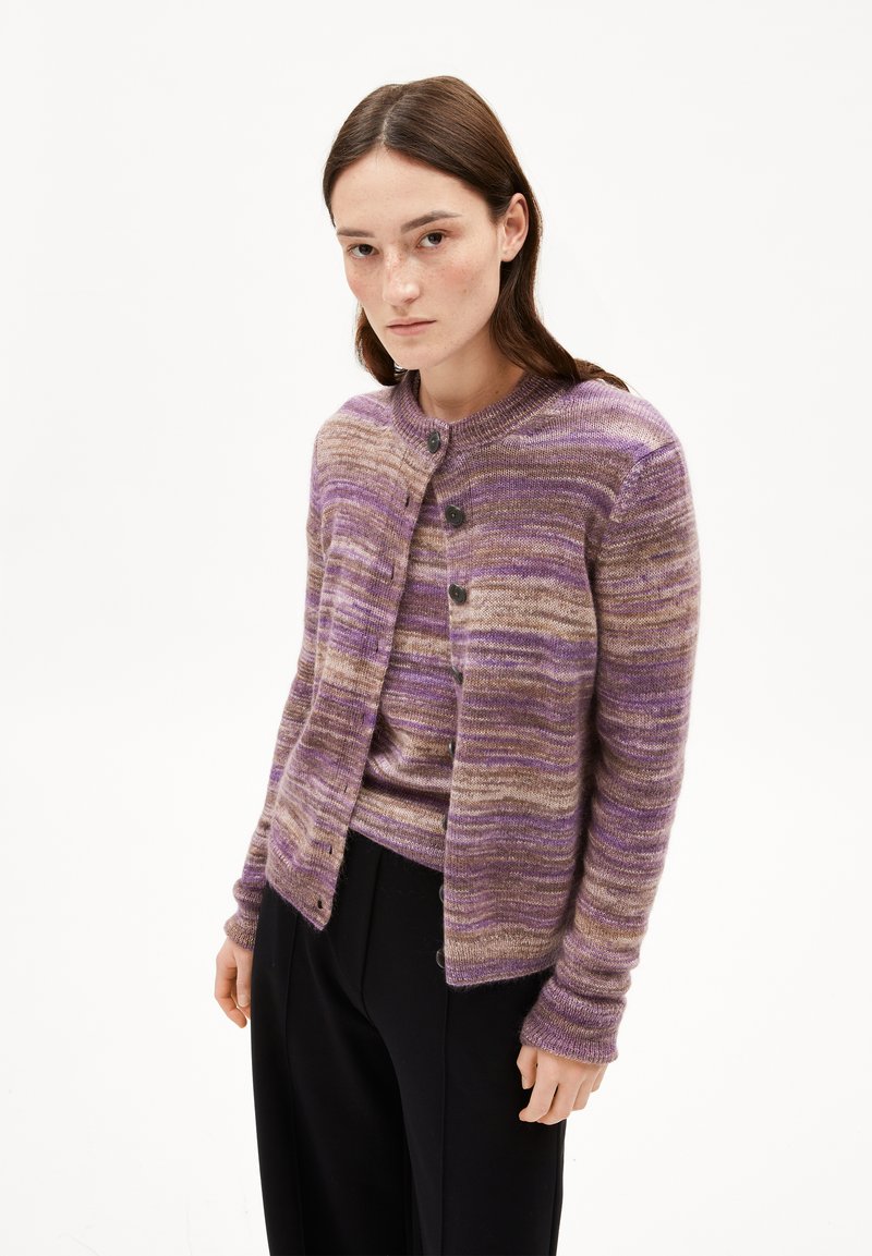 Femme portant un cardigan en tricot rayé violet et marron sur un haut assorti, associé à un pantalon noir, se tenant devant un fond blanc.