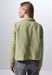 Chaqueta de pana verde con cuello, diseño ajustado y acabado texturizado, vista desde la parte trasera con vaqueros gris claro.