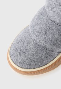 Pantufla gris de felpa con suela de goma, punta redondeada y ribete crema. Presenta detalles de costura y una superficie suave y texturizada.