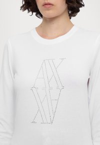 Camicia bianca a maniche lunghe caratterizzata da un design geometrico in strass, con "ARMANI EXCHANGE" stampato sotto in grigio. Materiale in cotone morbido.