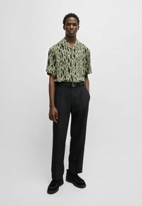 Kurzarmhemd mit grünem und schwarzem Schlangen-Muster, kombiniert mit hoch-taillierten schwarzen Hosen, getragen mit schwarzen Schuhen und minimalem Zubehör.