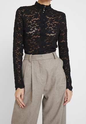 Blouse noire en dentelle florale à manches longues avec col montant et fermeture à boutons sur le devant, associée à un pantalon beige plissé taille haute.