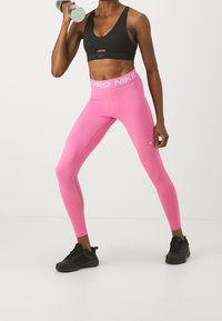 Svart sport-bh med en frontöppning och rosa högmidjade leggings med en vit Nike-logotyp. Svarta löparskor fulländar outfiten.