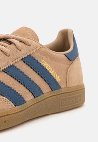 Béžová semišová teniska s modrými koženými pruhy, žlutým nápisem "SPEZIAL" a strukturovanou podrážkou z gumy. Má klasický design se třemi pruhy.