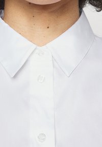Chemise blanche à boutons boutonnés avec un col traditionnel, une texture lisse, et des boutons en plastique transparent le long de la patte de boutonnage avant.