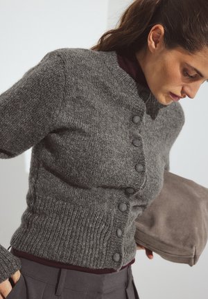 Grijze wollen cardigan met een geribbelde zoom, ronde halslijn en sluiting met zes knopen. Lange mouwen en zachte textuur, gedragen over een donker shirt.