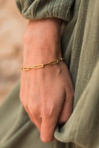 Zlatna narukvica s duguljastim ovalnim karikama, prikazana na ruci nasuprot maslinasto zelenoj tkanini. Glatka tekstura, minimalistički dizajn.