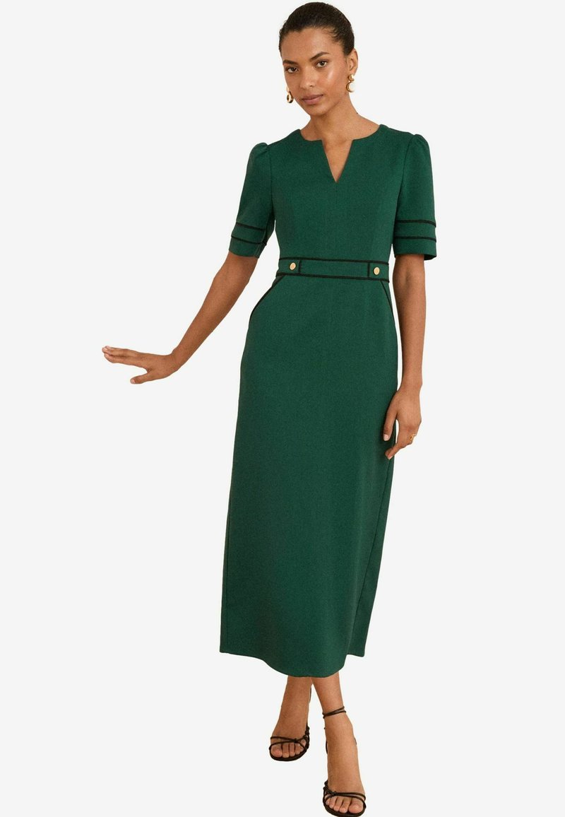 Femme portant une robe midi vert foncé avec de courtes manches bouffantes, un décolleté en V, des finitions noires et des détails en boutons dorés, associée à des talons noirs à lanières.
