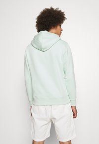 Jovem com cabelo cacheado a usar um hoodie verde menta claro e calções de carga brancos, de costas em relação a um fundo simples.