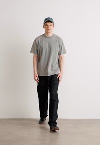 Gray cotton t-shirt with short sleeves, paired with black denim pants. The model wears a blue cap and black sneakers. Minimalist design.