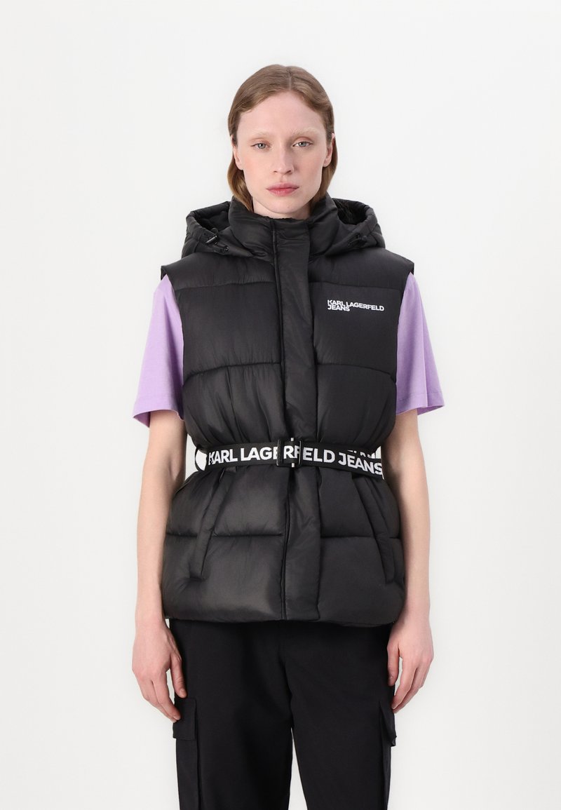 Juodas pūkuotas liemenė su kapišonu, su diržu su "KARL LAGERFELD JEANS" logotipu, vilkima ant alyvų spalvos marškinėlių. Lygus paviršius ir kvadratinė forma.