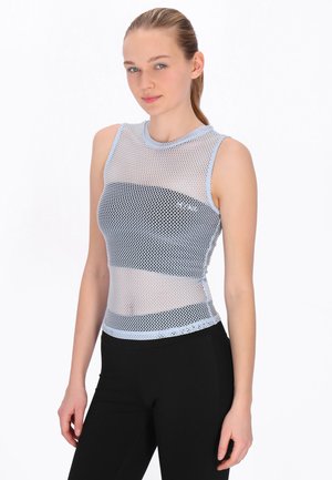 Femeie tânără cu păr blond, purtând un top alb fără mâneci din plasă peste un bandeau negru, asortat cu pantaloni negri, stând într-un cadru alb.