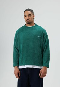 Groen, getextureerd fleece sweater met een ronde hals en lange mouwen, gecombineerd met een wit T-shirt. Minimale merkaanduiding op de borst.