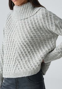 Femme portant un pull à col roulé gris clair en maille torsadée avec des manches longues et un pantalon taille haute foncé, main posée sur la hanche.