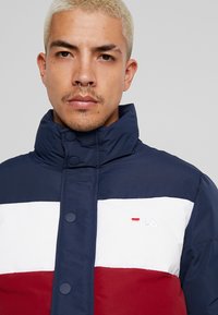 Veste rembourrée avec un col haut, en blocs de couleurs marine, blanche et rouge. Logo brodé sur la poitrine avec des boutons-pression pour la fermeture.