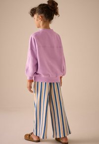 Fialová oversized mikina s balónovými rukávy, kombinovaná s širokými pruhovanými kalhotami v béžové a modré barvě. Outfit doplňují hnědé nazouvací boty.
