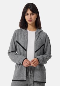 Grå zip-up hoodie i mjukt tyg, med svarta accenter, huva och en broderad logotyp på bröstet. Händerna visas sammanflätade.