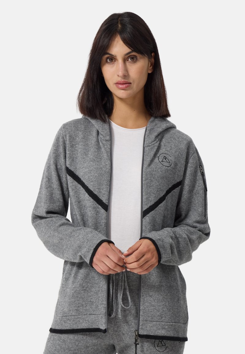 Grå zip-up hoodie i mjukt tyg, med svarta accenter, huva och en broderad logotyp på bröstet. Händerna visas sammanflätade.