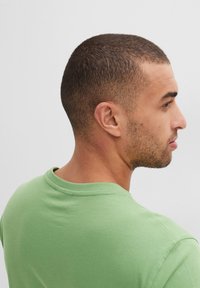 T-shirt in cotone verde con scollatura rotonda e maniche corte, che mostra una texture liscia e un design semplice. Nessun motivo o accessorio visibile.