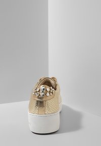 Zapatilla texturizada en oro con suela plataforma blanca, adornada con grandes rhinestones transparentes en la lengüeta del talón, vista desde la parte posterior.