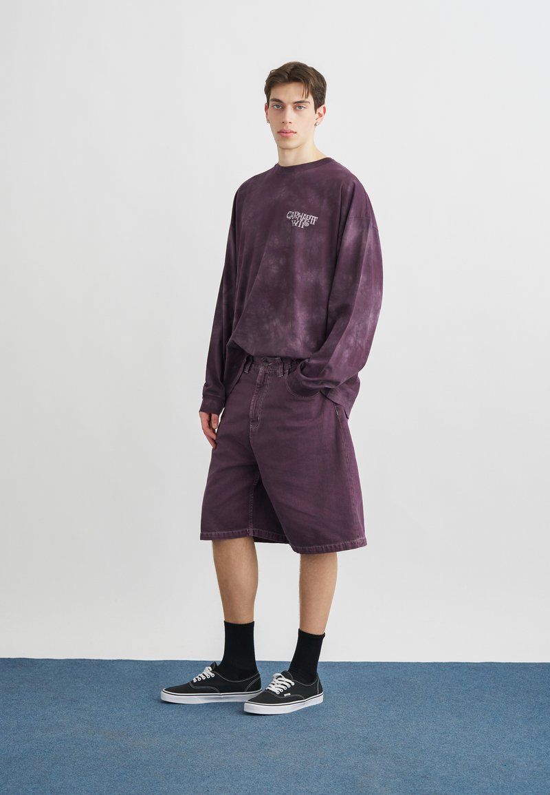 Giovane in piedi che indossa una maglia a maniche lunghe viola con effetto tie-dye, pantaloncini abbinati fino al ginocchio, calze nere e sneakers basse nere.