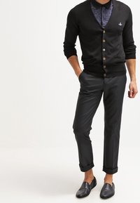 Černý cardigan s knoflíky přes tmavě vzorovanou košili, v kombinaci s dobře střiženými černými kalhotami a černými slip-on botami.
