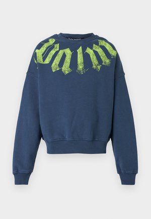 Homme portant des lunettes de soleil foncées, un sweat-shirt bleu marine avec un texte vert, un short en jean, des chaussettes blanches et des baskets vertes avec des lacets orange et bordeaux.