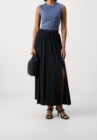 Žena v bezrukávovém modrém topu, černé maxi sukni s vysokým pasem a bočním rozparkem, černých sandálech na podpatku a držící černou kabelku.