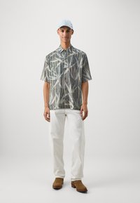 Short-sleeve shirt with a green and beige bamboo pattern, paired with white trousers and brown boots, complemented by a light blue cap.