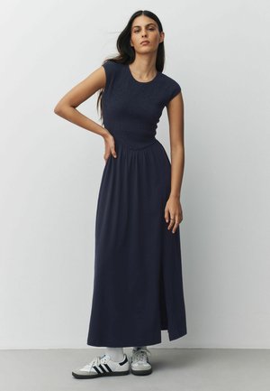 Kvinde iført en mørkeblå ærmeløs maxikjole med en tekstureret overdel og hvide sneakers, stående med den ene hånd på hoften.