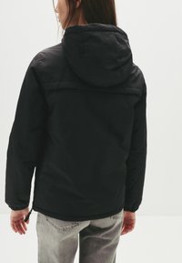 Sort hættejakke med en glat overflade, der har en lynlåslukning og elastiske manchetter, parret med lysegrå jeans.