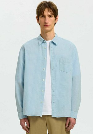 Junger Mann, der ein hellblaues Hemd über einem weißen T-Shirt und beige Hose trägt, vor schlichtem Hintergrund stehend.