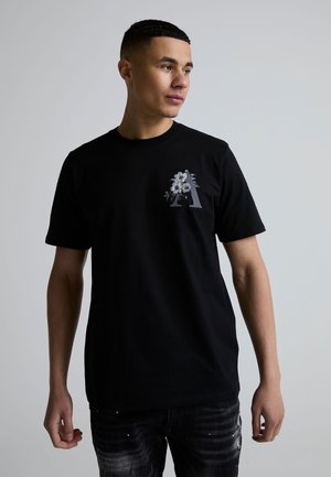 Ung mand iført en sort t-shirt med et blomsterdesign i form af bogstavet "A", stående mod en ensfarvet lysegrå baggrund og kigger mod højre.