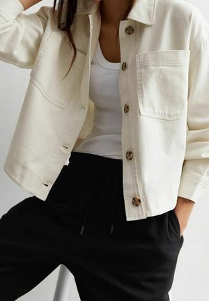 Henkilö pukeutuneena maitokermanväriseen napilliseen takkiin valkoisen toppiin ja mustiin narulla kiristettäviin housuihin, toinen käsi taskussa.