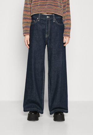 Osoba ubrana w ciemnoniebieskie dżinsy o szerokich nogawkach, czarne buty na platformie oraz wielokolorowy sweter w paski, stojąca na jednolitym tle.