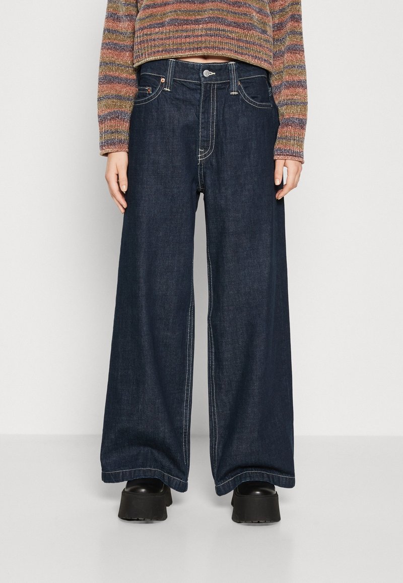 Personne portant un jean pattes d'éléphant bleu foncé, des chaussures à plateforme noires et un pull rayé multicolore, debout devant un fond uni.