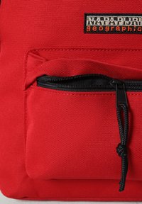 Roter Stoffrucksack mit einer vorderen Reißverschlusstasche, schwarzem Reißverschluss und Markenschild. Das Material scheint strukturiert und langlebig zu sein, mit einem schlichten Design.