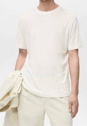 Homme portant un t-shirt à manches courtes blanc cassé uni et un pantalon, tenant une veste assortie blanc cassé dans la main gauche.