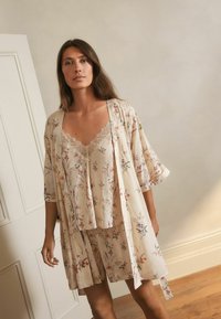 Cvjetni kratki set od mekog bež materijala s čipkastim obrubom. Sadrži labavi kimono stil pokrivača i elastičnu traku u struku, prikazujući delikatne uzorke.